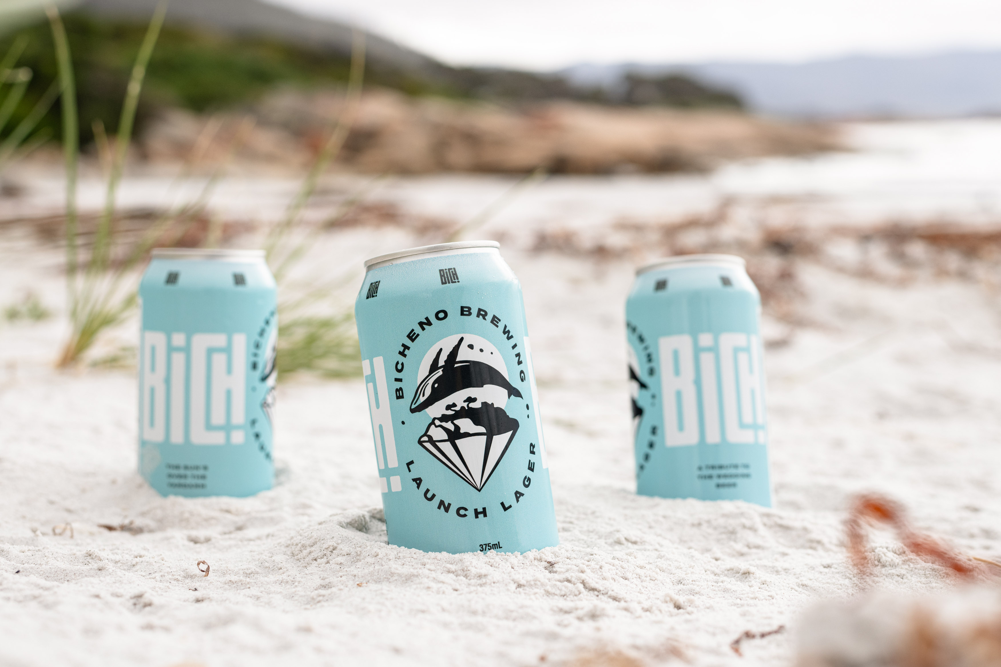 Three Blue Bicheno Brewing cans sitting in the sand.